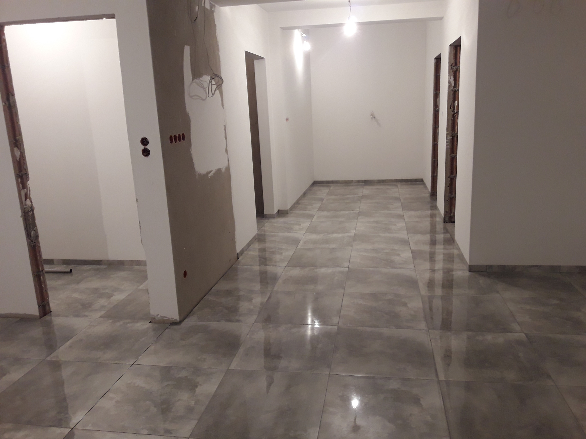 Wykończone płytki podłogowe w korytarzu, widoczne przygotowania do malowania ścian i montażu drzwi. Szare płytki z połyskiem, surowy stan wykończenia.