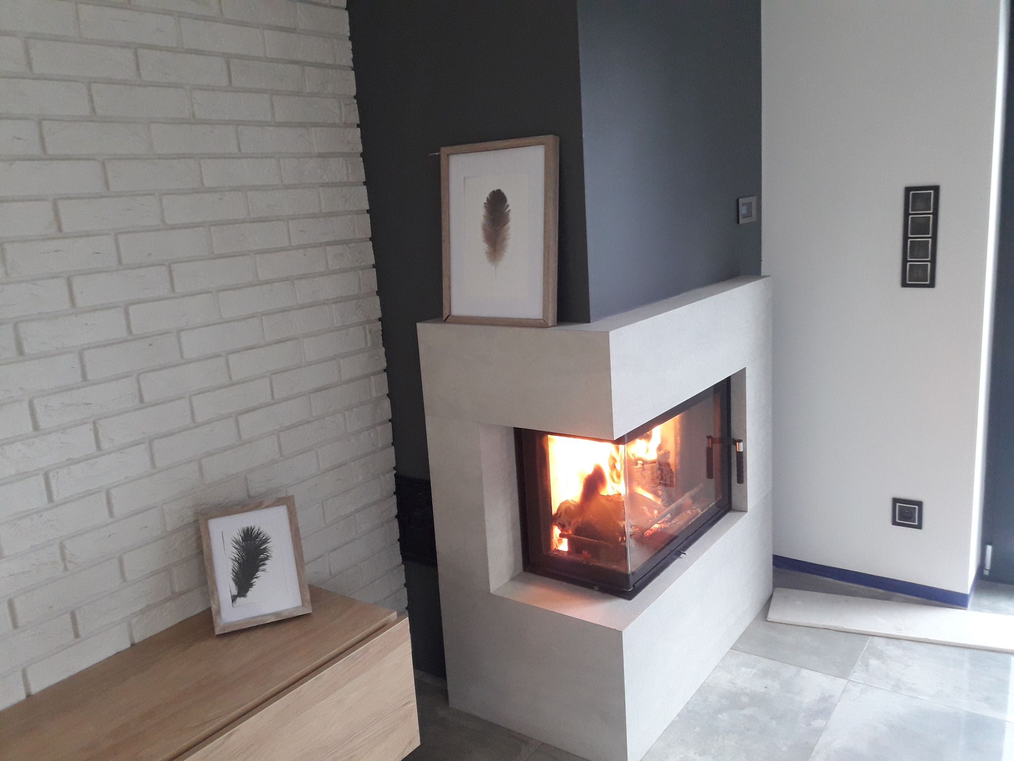 Nowoczesny kominek z paleniskiem w salonie w Zdunach. Biała cegła na ścianie, minimalistyczny design, płomienie widoczne przez szybę. Element wykończenia wnętrza.