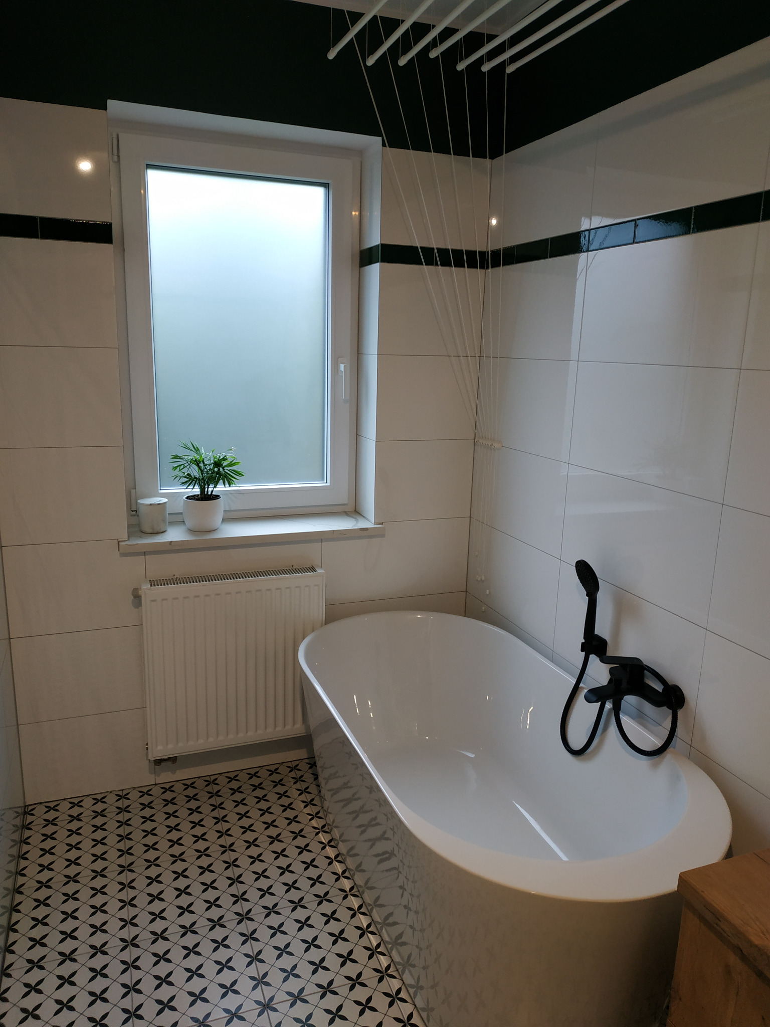 Biała wanna z czarną armaturą w nowoczesnej łazience z biało-czarną mozaiką na podłodze i rośliną w oknie. Minimalistyczny design z akcentami kolorystycznymi.