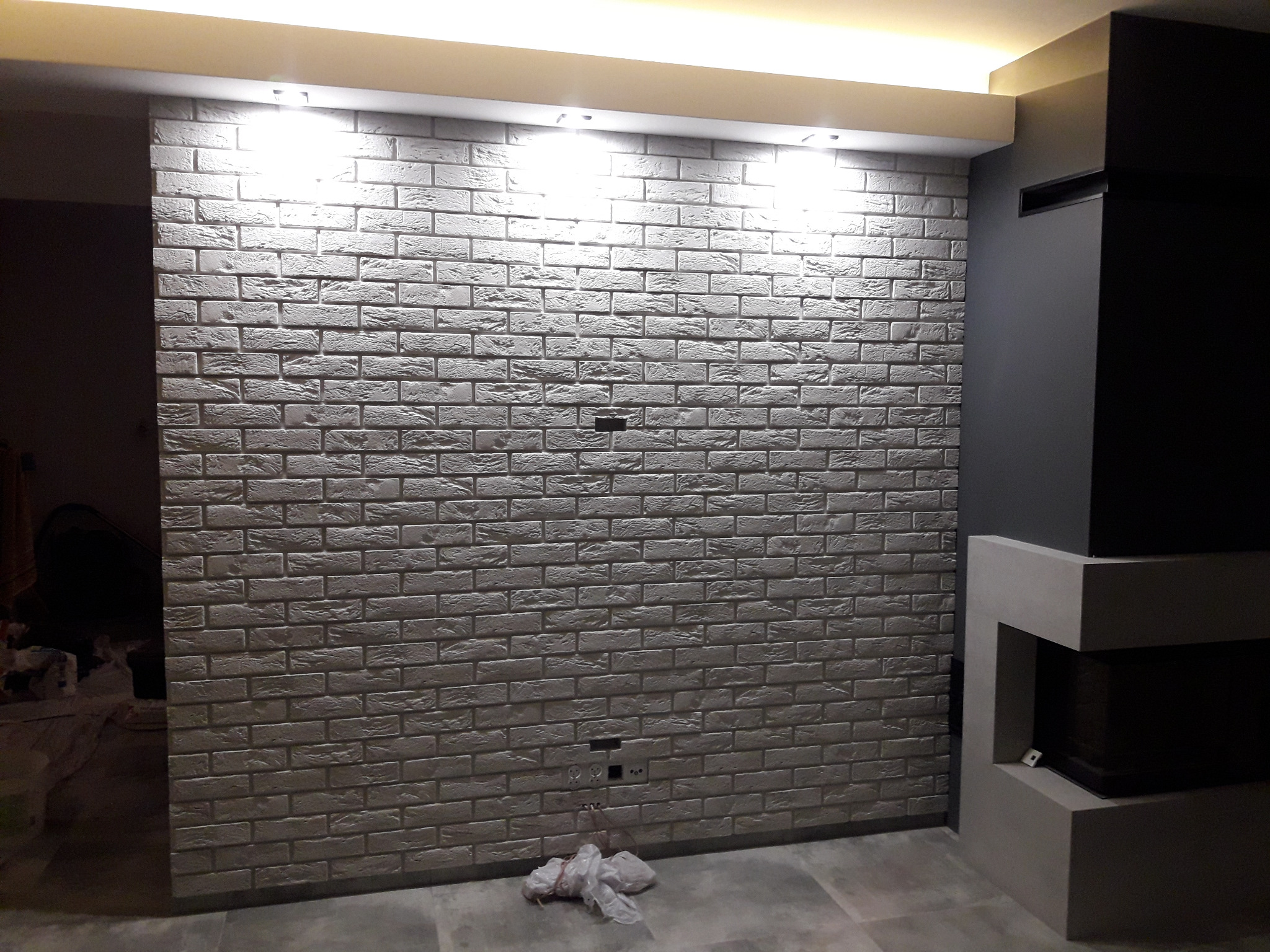 Ściana z dekoracyjnej cegły w szarościach, oświetlona punktowo, z fragmentem nowoczesnego kominka po prawej stronie. Widoczne gniazdka elektryczne i fragment podłogi.
