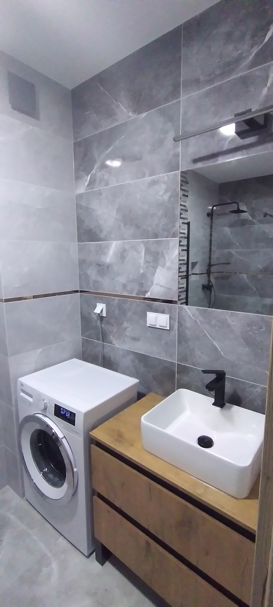 Wykończona łazienka w szarościach z marmurkowymi płytkami, białą umywalką nablatową na drewnianej szafce i pralką. Widoczne detale: czarna bateria, lustro.