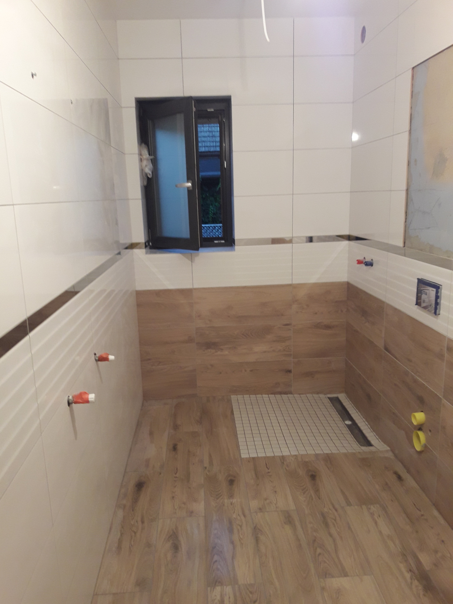 Ukończona łazienka z drewnopodobnymi i białymi płytkami, widoczny odpływ liniowy w kabinie prysznicowej, okno z czarną ramą i wystające zawory.