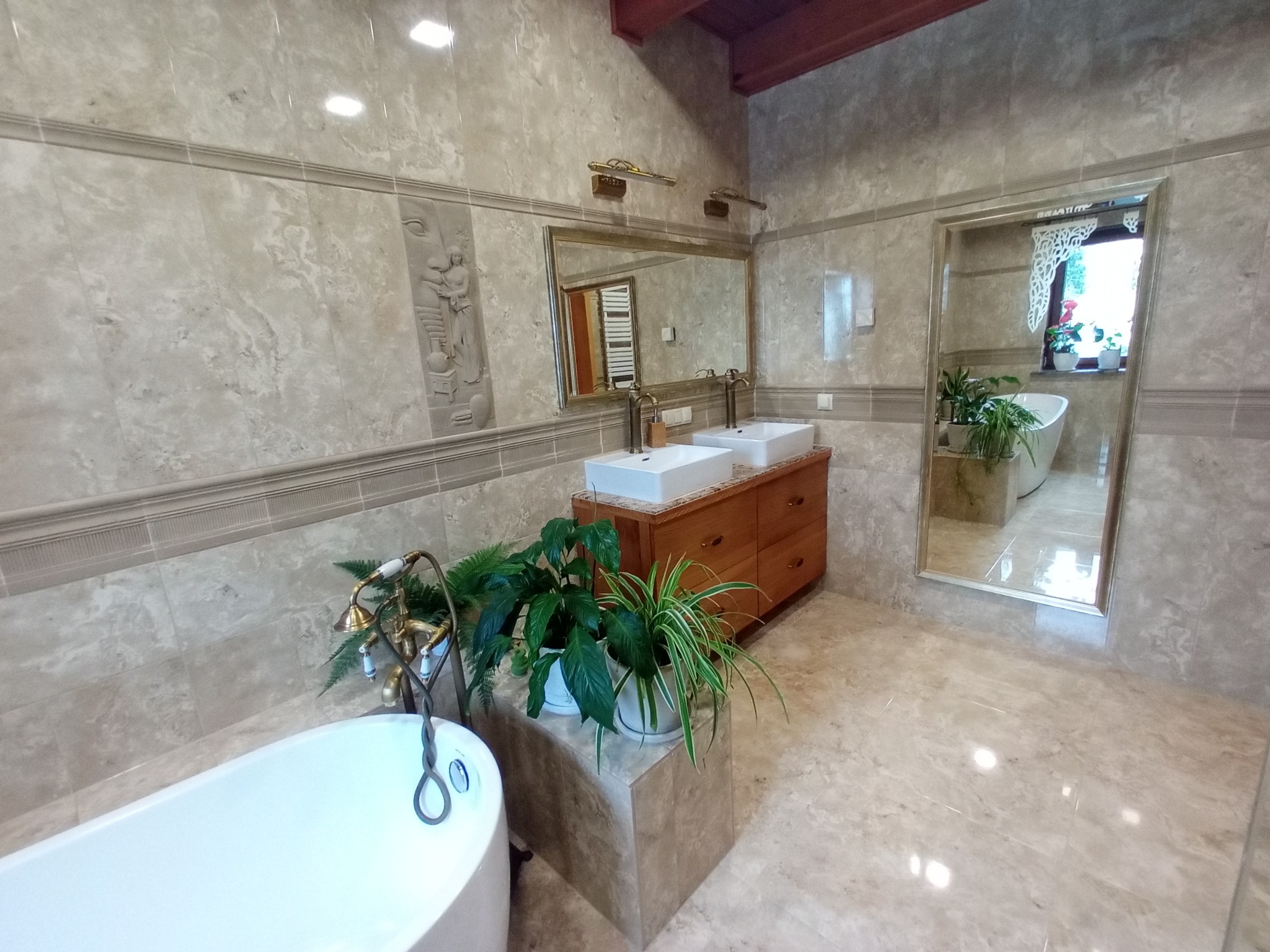 Elegancka łazienka w beżowych płytkach z dekoracyjnym reliefem, drewnianą szafką pod umywalki i roślinami doniczkowymi, odbicie w lustrze dodaje głębi.