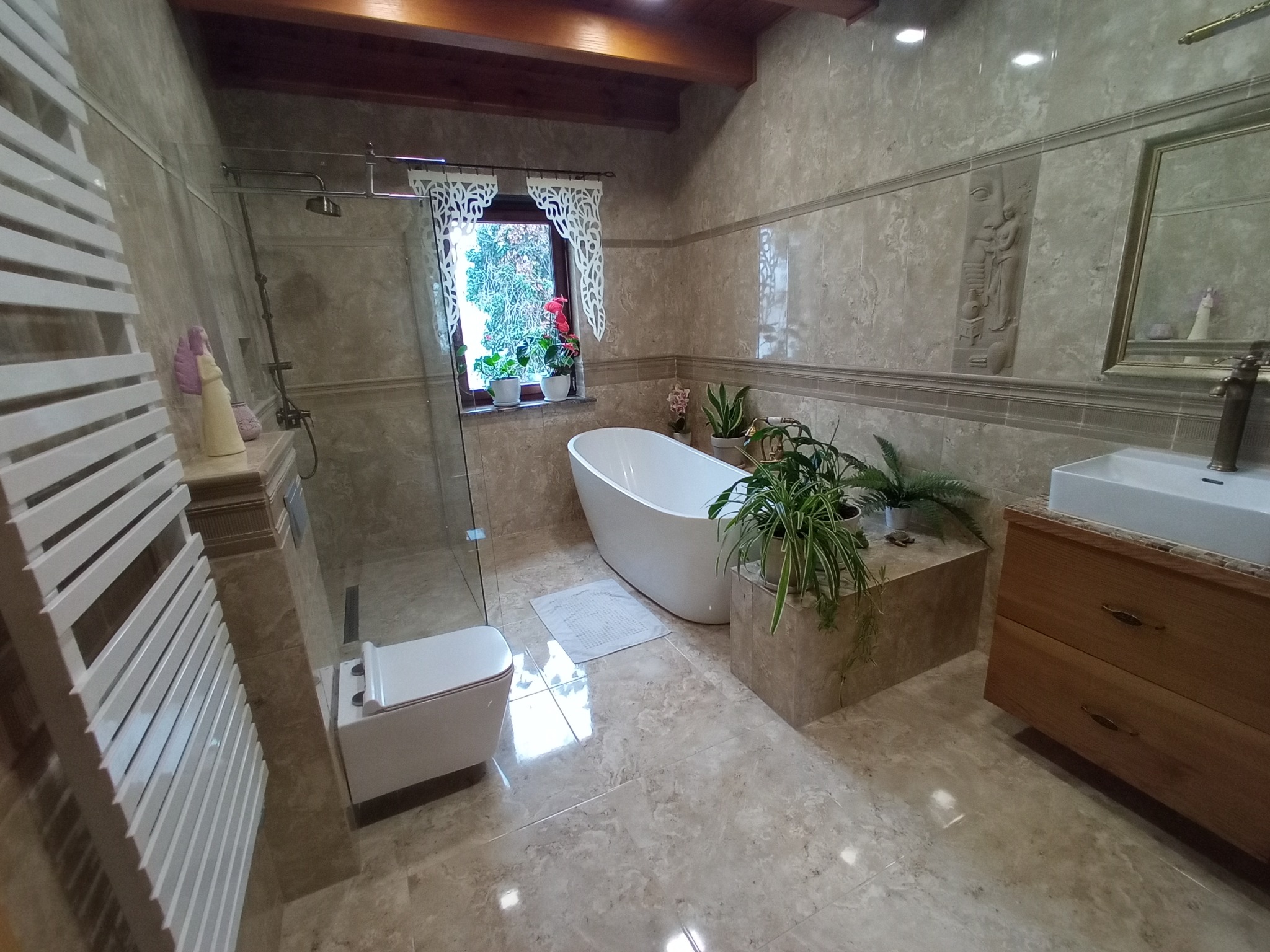 Elegancka łazienka w beżowych płytkach z wolnostojącą wanną, kabiną prysznicową i drewnianą szafką pod umywalką. Okno zdobią białe, dekoracyjne firanki.