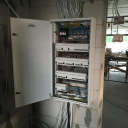 GRENOVA Marcin Gregorczuk - Otwarta skrzynka elektryczna z widocznymi bezpiecznikami i przewodami, zamontowana na ścianie z betonu komórkowego w surowym wnętrzu.