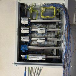 GRENOVA Marcin Gregorczuk - Rozdzielnica elektryczna w szarej obudowie z widocznymi bezpiecznikami, przewodami w kolorach: niebieskim, żółtym i zielonym. Instalacja w trakcie montażu.