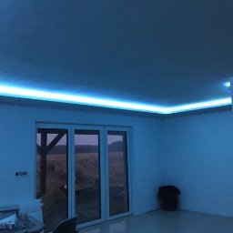 GRENOVA Marcin Gregorczuk - Nowoczesne oświetlenie LED wzdłuż sufitu, tworzące niebieską poświatę w jasnym wnętrzu z dużym oknem balkonowym i widokiem na zewnątrz. Minimalistyczny design.