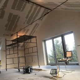 GRENOVA Marcin Gregorczuk - Poddasze w trakcie wykończenia: rusztowanie, płyty gipsowe na skosach, lampa budowlana. Widoczne okno z widokiem na zieleń. Krzesło i narzędzia sugerują przerwę w pracy.