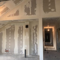 GRENOVA Marcin Gregorczuk - Wnętrze w trakcie szpachlowania ścian gipsowych, widoczne niedoskonałości i przygotowanie do malowania. W tle korytarz z widocznymi drzwiami i oświetleniem.