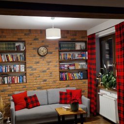 GRENOVA Marcin Gregorczuk - Salon z ceglaną ścianą, drewnianymi półkami z książkami i szarą sofą z czerwonymi poduszkami w kratę. Okno z czerwonymi zasłonami w kratę. Ciepłe oświetlenie.