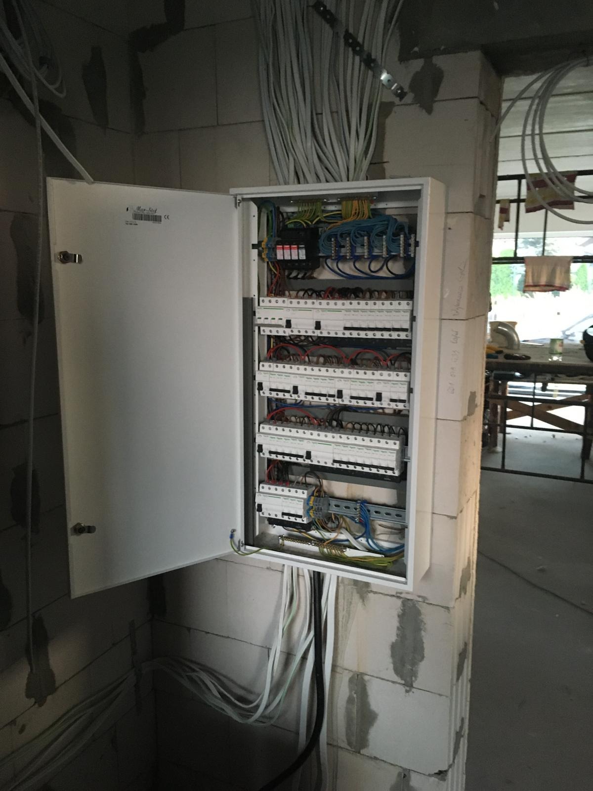 Otwarta skrzynka elektryczna z widocznymi bezpiecznikami i przewodami, zamontowana na ścianie z betonu komórkowego w surowym wnętrzu.