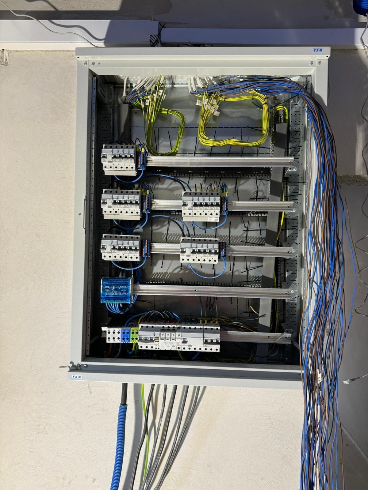 Rozdzielnica elektryczna w szarej obudowie z widocznymi bezpiecznikami, przewodami w kolorach: niebieskim, żółtym i zielonym. Instalacja w trakcie montażu.