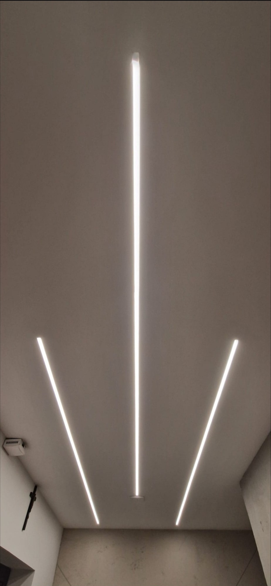 Nowoczesne oświetlenie sufitowe z trzema liniowymi lampami LED, tworzącymi geometryczny wzór. Minimalistyczny design, ciepłe światło, widoczny fragment ściany i sufitu.