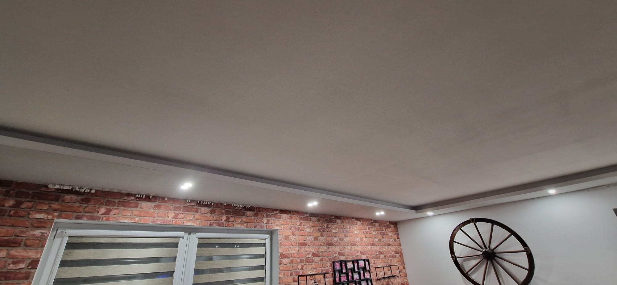 Wykończone wnętrze z ceglaną ścianą, oknem z żaluzjami i dekoracyjnym kołem na ścianie. Sufit podwieszany z oświetleniem punktowym. Widok z dołu.