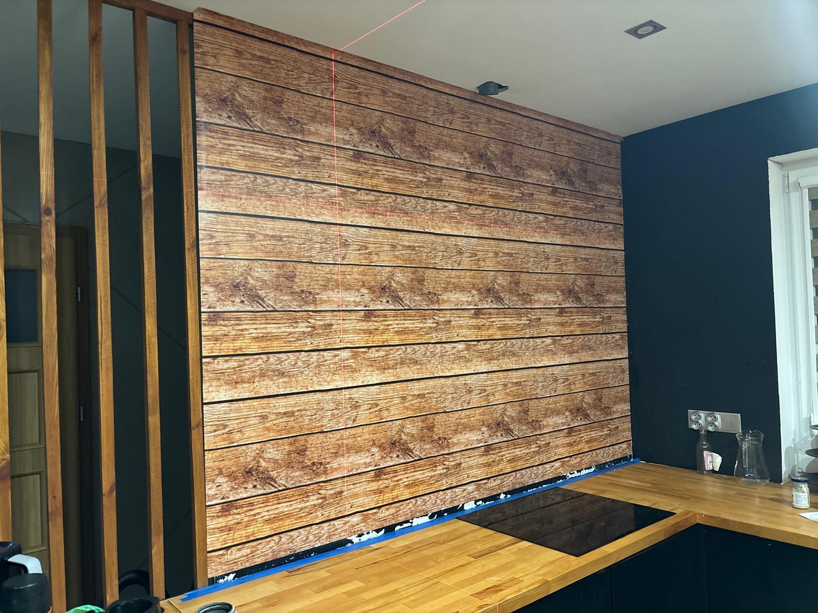 Ściana wykończona panelami imitującymi drewno, z widocznym laserowym poziomowaniem, fragmentem sufitu z oświetleniem i elementami zabudowy kuchennej w tle.