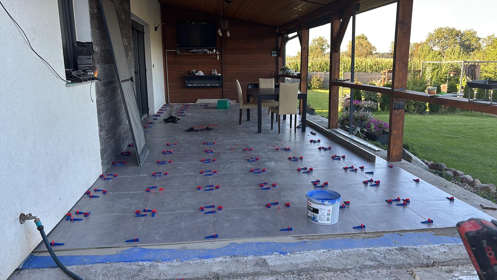 Montaż płytek na tarasie z użyciem systemu poziomowania. Widoczne płytki, krzyżyki dystansowe, wiadro z klejem i narzędzia. Częściowo ukończony taras.