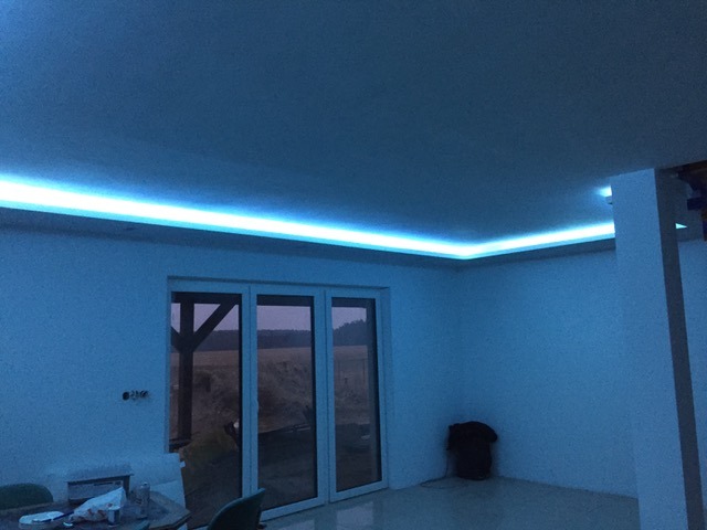 Nowoczesne oświetlenie LED wzdłuż sufitu, tworzące niebieską poświatę w jasnym wnętrzu z dużym oknem balkonowym i widokiem na zewnątrz. Minimalistyczny design.