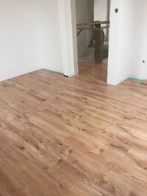 Ułożona podłoga z paneli drewnopodobnych w jasnym wnętrzu. Widoczne białe ściany i fragment przejścia do innego pomieszczenia w trakcie remontu.