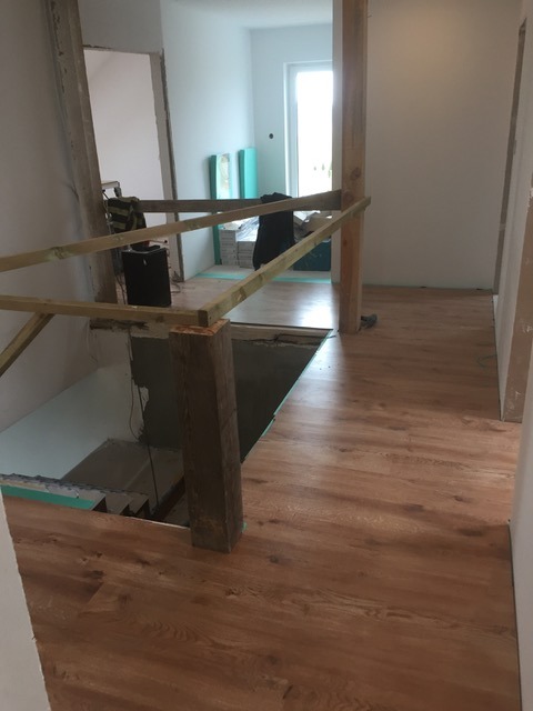 Układanie paneli podłogowych w jasnym wnętrzu domu, widoczne drewniane słupy i fragment schodów w trakcie remontu. Częściowo wykończona podłoga.