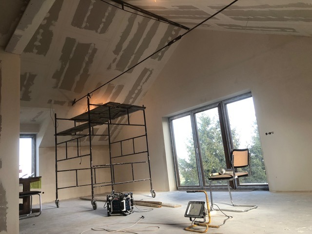 Poddasze w trakcie wykończenia: rusztowanie, płyty gipsowe na skosach, lampa budowlana. Widoczne okno z widokiem na zieleń. Krzesło i narzędzia sugerują przerwę w pracy.