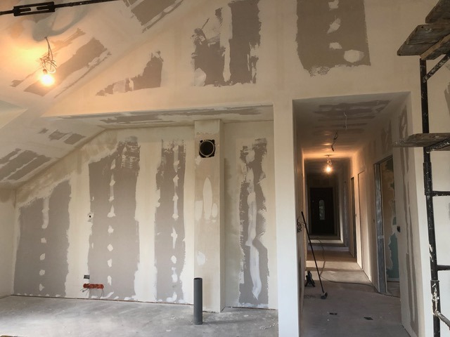 Wnętrze w trakcie szpachlowania ścian gipsowych, widoczne niedoskonałości i przygotowanie do malowania. W tle korytarz z widocznymi drzwiami i oświetleniem.