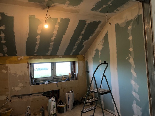 Poddasze w trakcie wykończenia: ściany z płyt gipsowo-kartonowych, częściowo zaszpachlowane, widoczne okno, drabina i worki z zaprawą. Oświetlenie żarówką na kablu.