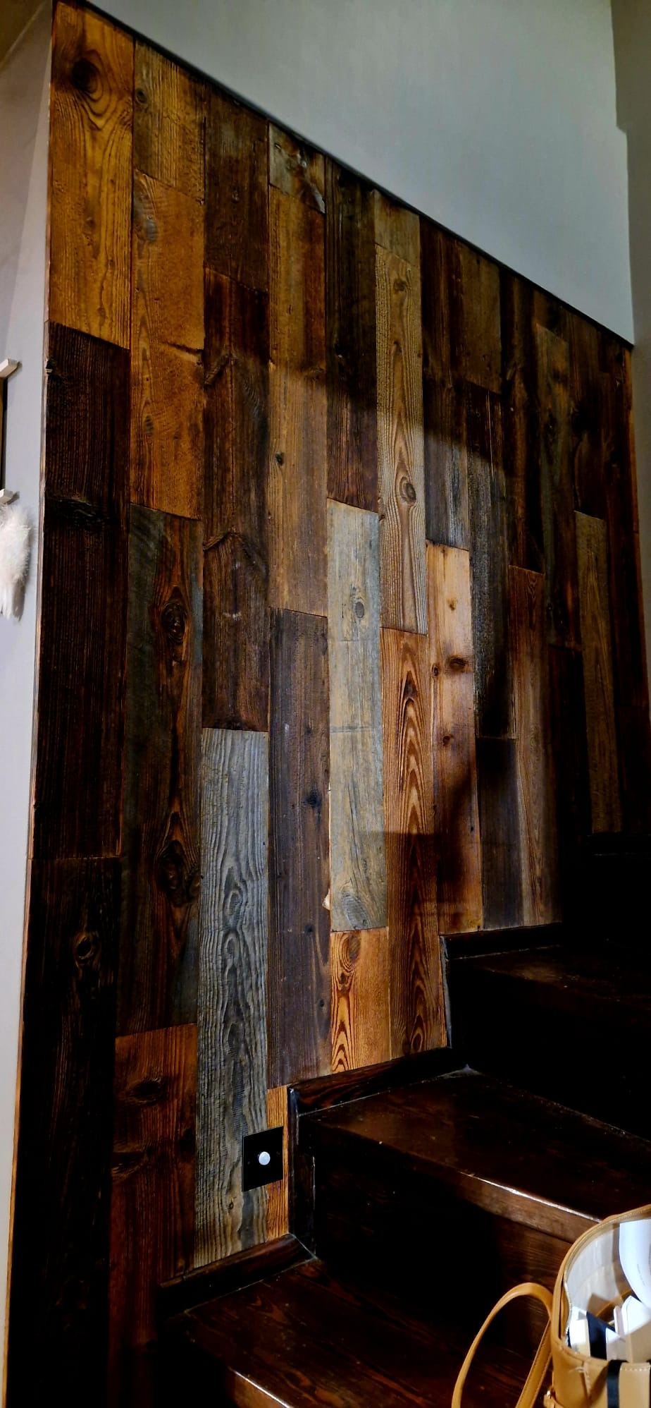 Ściana z desek o zróżnicowanej kolorystyce drewna, przylegająca do drewnianych schodów z podświetleniem LED. Dekoracyjne wykończenie wnętrza z naturalnych materiałów.