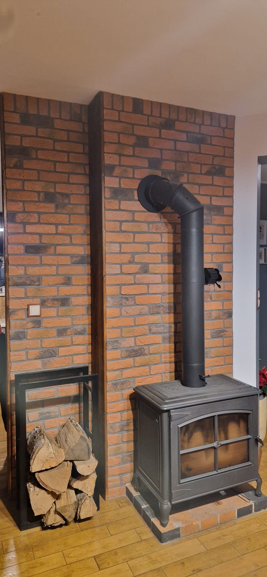 Kominek żeliwny z czarnym przewodem kominowym na tle ściany z cegły w Sepno. Obok stojak na drewno. Podłoga wyłożona panelami imitującymi drewno.