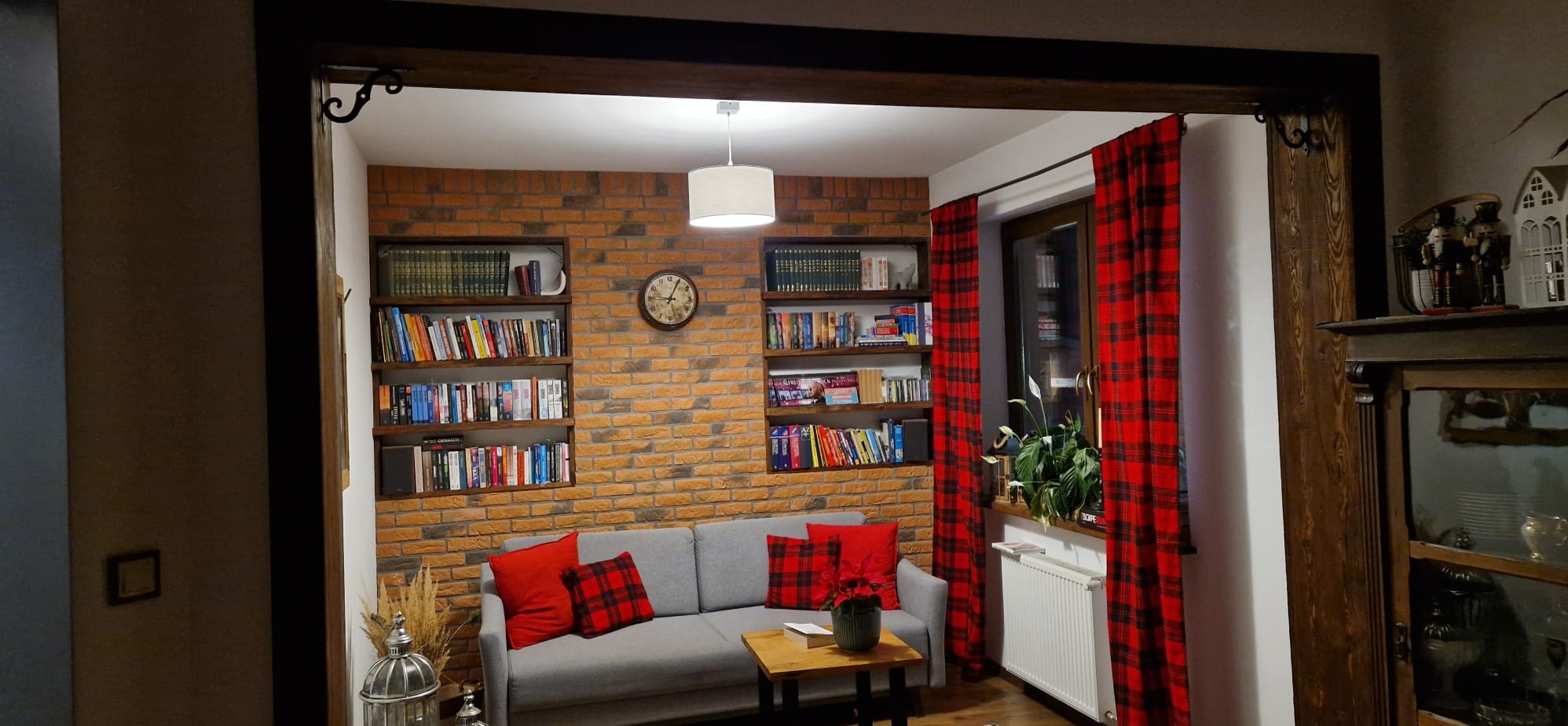 Salon z ceglaną ścianą, drewnianymi półkami z książkami i szarą sofą z czerwonymi poduszkami w kratę. Okno z czerwonymi zasłonami w kratę. Ciepłe oświetlenie.