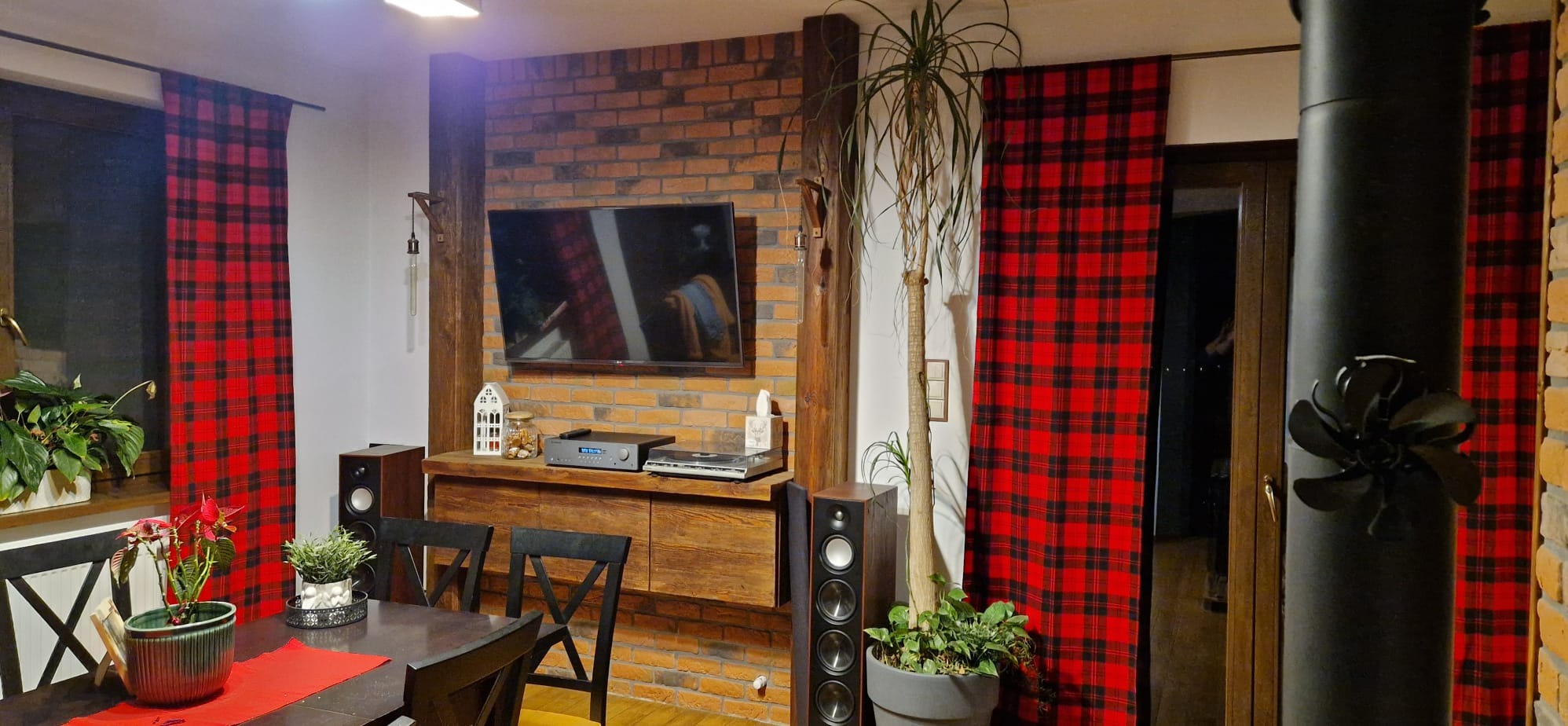 Salon z ceglaną ścianą, drewnianymi elementami i czerwono-czarnymi zasłonami w kratę. Widoczny telewizor, sprzęt audio i rośliny doniczkowe. Styl rustykalny.
