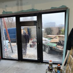 FENSKE INVESTMENTS GROUP SPÓŁKA Z OGRANICZONĄ ODPOWIEDZIALNOŚCIĄ - Czarne, trzyskrzydłowe okno z widokiem na podwórze podczas montażu. Widoczne narzędzia, drabina i materiały budowlane. Ściany w trakcie wykończenia.