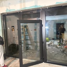 FENSKE INVESTMENTS GROUP SPÓŁKA Z OGRANICZONĄ ODPOWIEDZIALNOŚCIĄ - Czarne, panoramiczne okno z otwartym skrzydłem w surowym wnętrzu. Widoczne narzędzia i osoba w tle, co sugeruje etap montażu. Charakterystyczne mocowania na górze ramy.