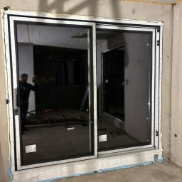 FENSKE INVESTMENTS GROUP SPÓŁKA Z OGRANICZONĄ ODPOWIEDZIALNOŚCIĄ - Zamontowane, czarne drzwi przesuwne z białymi ramami, widoczne pianka montażowa i surowy beton. W tle widoczny pracownik i wnętrze w trakcie wykończenia.