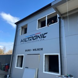 FENSKE INVESTMENTS GROUP SPÓŁKA Z OGRANICZONĄ ODPOWIEDZIALNOŚCIĄ - Szara hala serwisu samochodowego z napisem 'WILTRONIC' i oknami w trakcie montażu. Widoczny pracownik w oknie na piętrze. Na dole paczka z oknami.