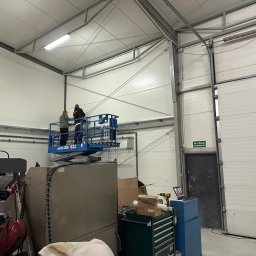 FENSKE INVESTMENTS GROUP SPÓŁKA Z OGRANICZONĄ ODPOWIEDZIALNOŚCIĄ - Montaż bramy garażowej segmentowej w hali produkcyjnej. Dwóch monterów na podnośniku koszowym przy suficie. Widoczne narzędzia i elementy konstrukcyjne bramy.