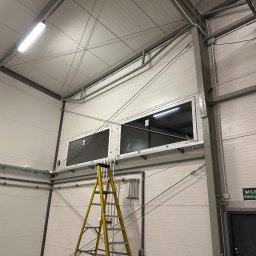 FENSKE INVESTMENTS GROUP SPÓŁKA Z OGRANICZONĄ ODPOWIEDZIALNOŚCIĄ - Dwa okna zamontowane wysoko w hali przemysłowej, widoczne z żółtej drabiny. Białe ściany z panelami, metalowa konstrukcja dachu i instalacje. Tabliczka 'Wyjście ewakuacyjne'.