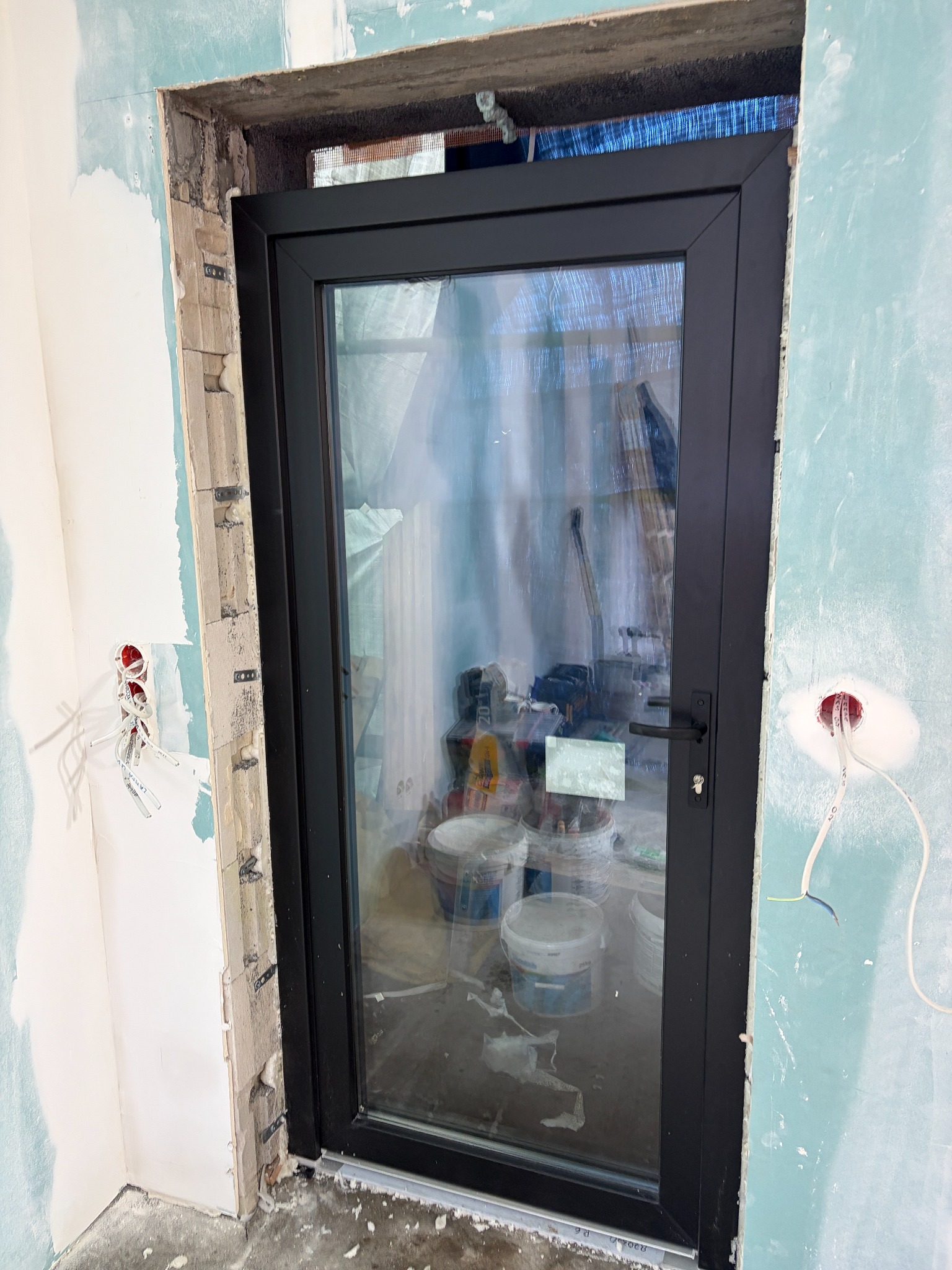 Czarne, szklane drzwi przesuwne zamontowane w surowym otworze ściany z widocznymi puszkami elektrycznymi i wiadrami farby w tle.