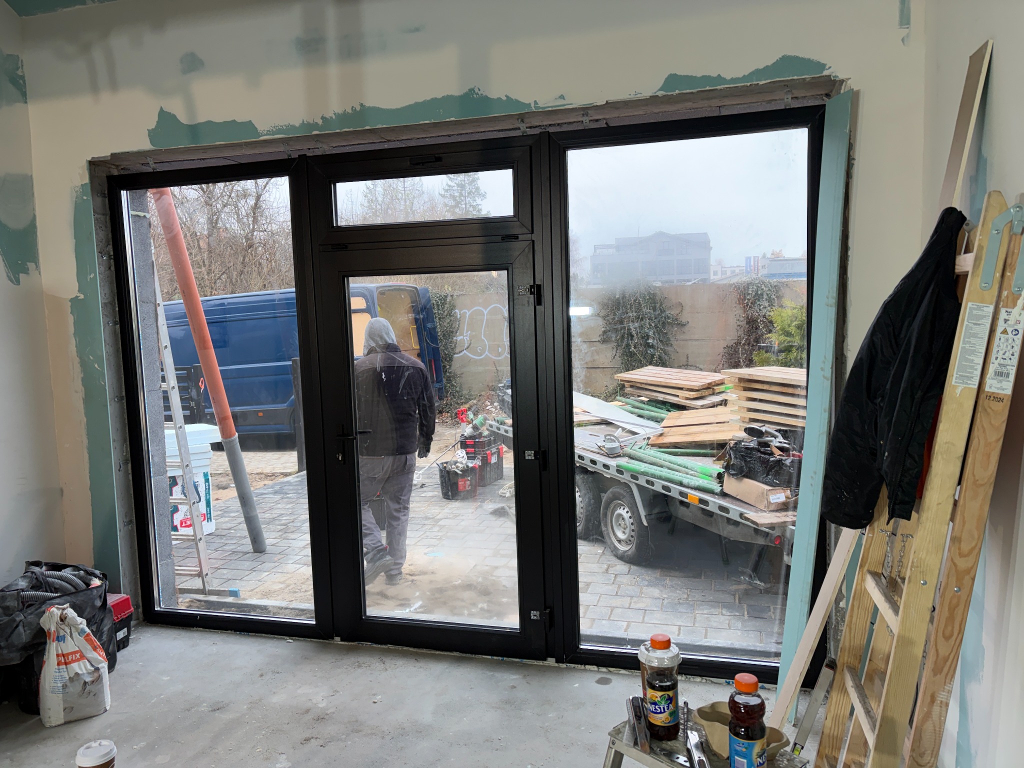 Czarne, trzyskrzydłowe okno z widokiem na podwórze podczas montażu. Widoczne narzędzia, drabina i materiały budowlane. Ściany w trakcie wykończenia.