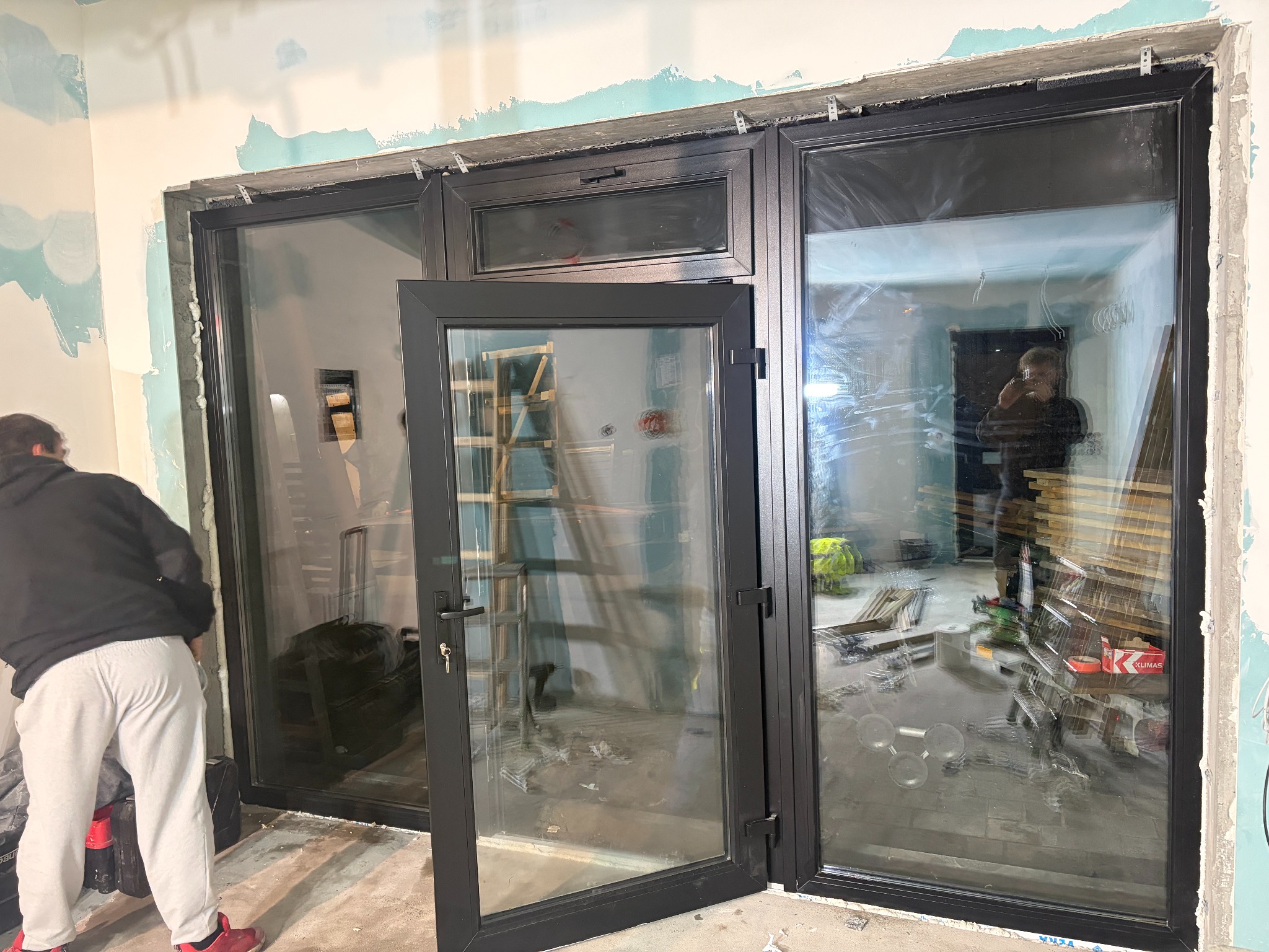 Czarne, panoramiczne okno z otwartym skrzydłem w surowym wnętrzu. Widoczne narzędzia i osoba w tle, co sugeruje etap montażu. Charakterystyczne mocowania na górze ramy.