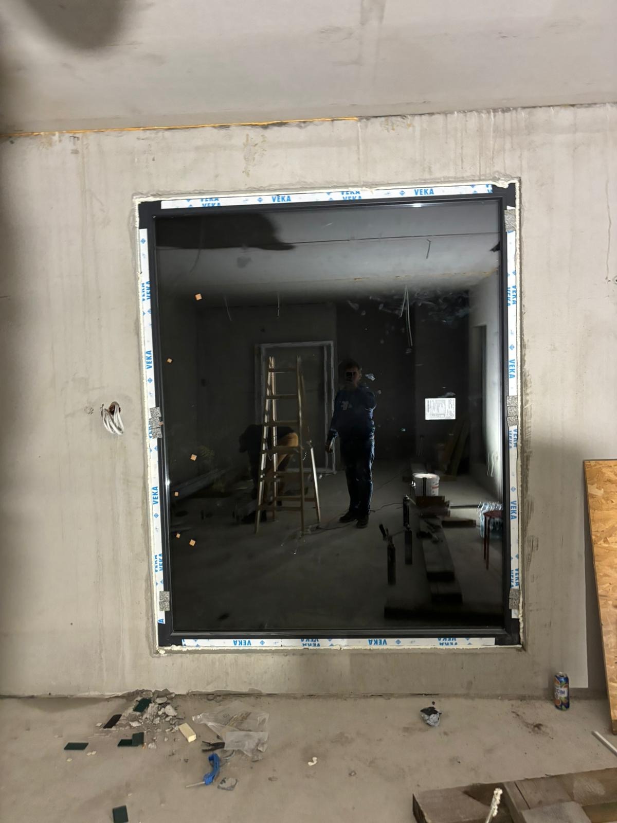 Zamontowane okno marki VEKA w surowym wnętrzu. Widoczne ślady prac budowlanych i odbicie osoby w szybie. Surowy beton na ścianach i podłodze.