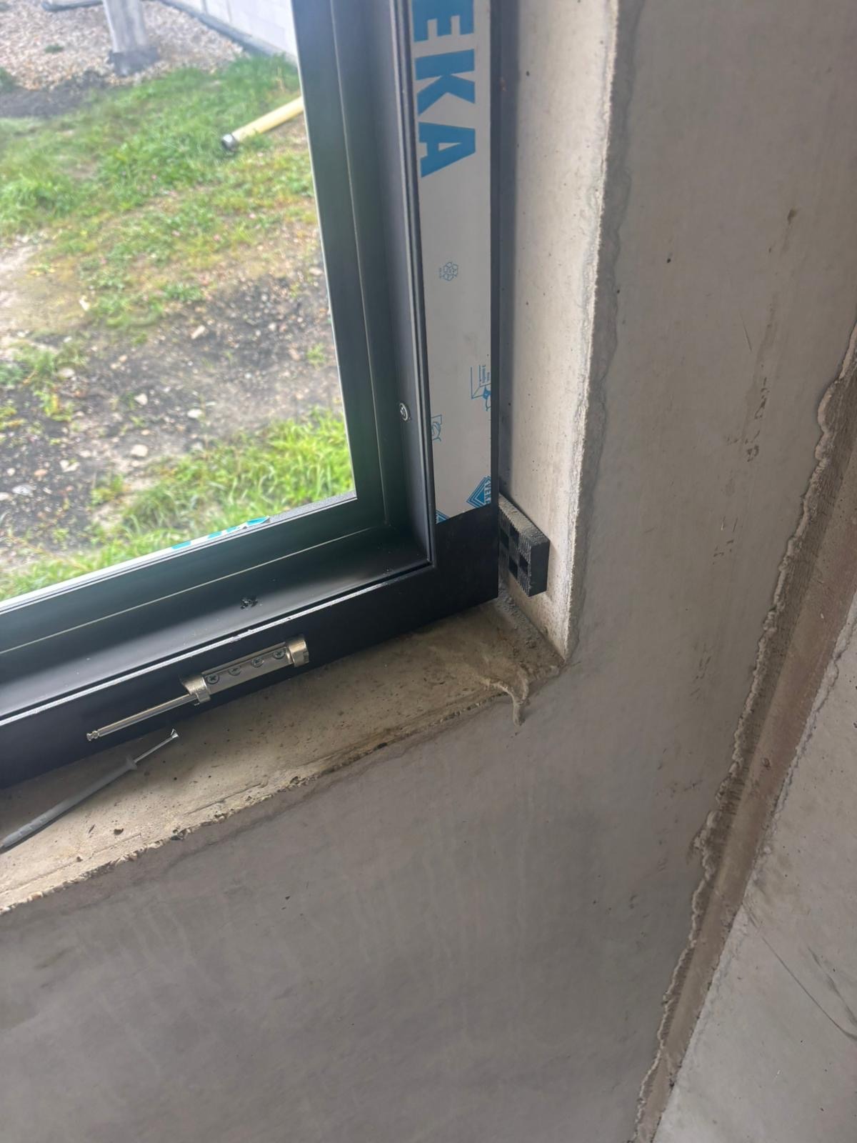 Ciemna rama okienna w surowym otworze ściennym, widoczne detale montażu i uszczelnienia. Widok na zewnątrz przez szybę.