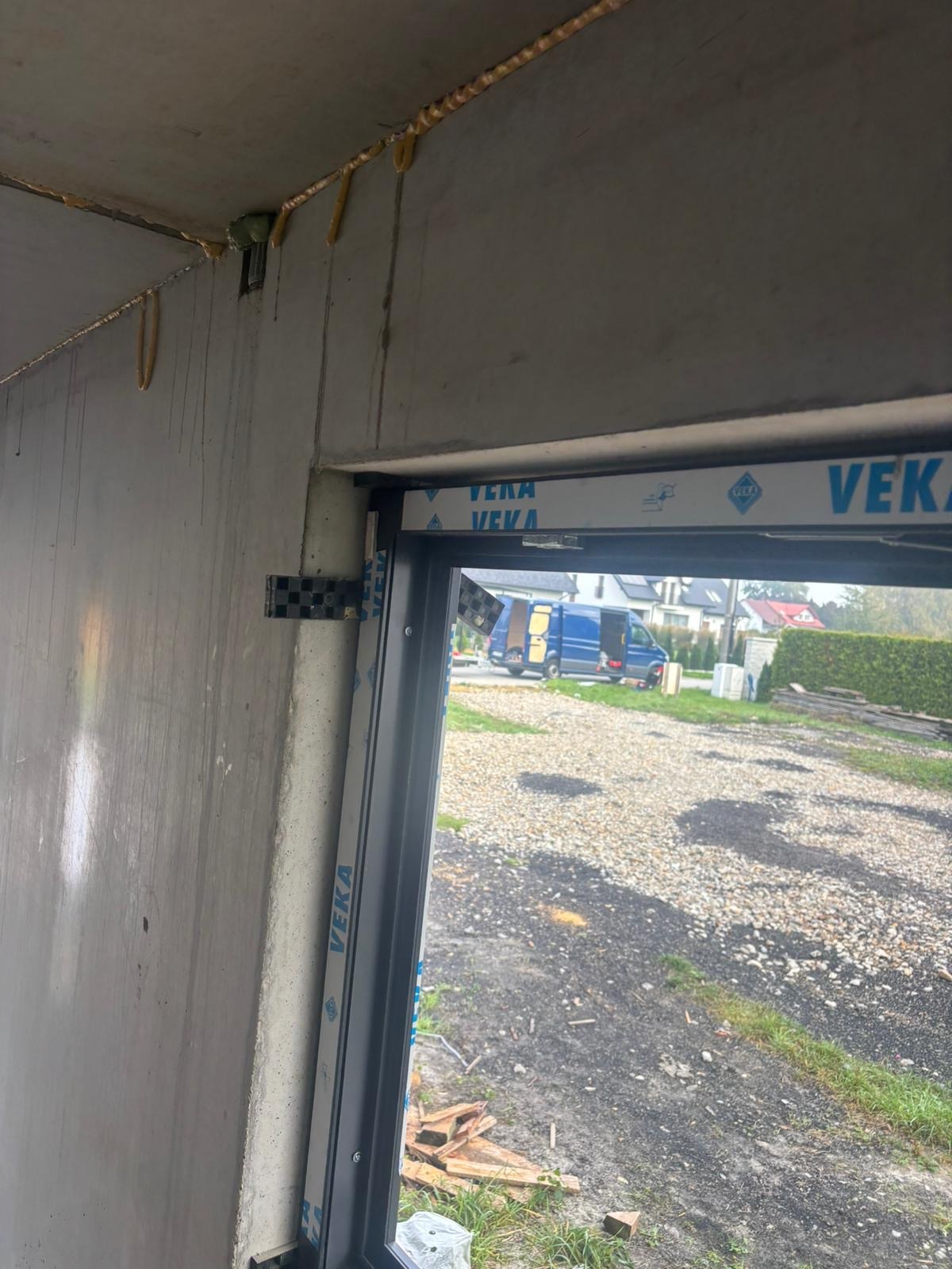 Surowy montaż okna marki VEKA w Warszawie, widok z wnętrza budynku na zewnątrz. Na zewnątrz zaparkowany niebieski bus. Widoczne pianki montażowe.