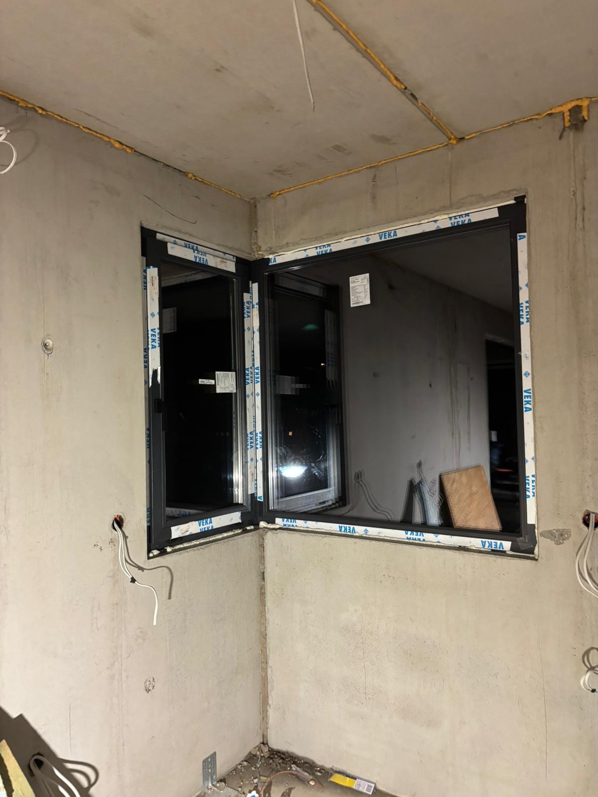 Montaż narożnego okna w surowym wnętrzu. Widoczne pianka montażowa, kable elektryczne i fragmenty materiałów budowlanych. Okno marki VEKA.