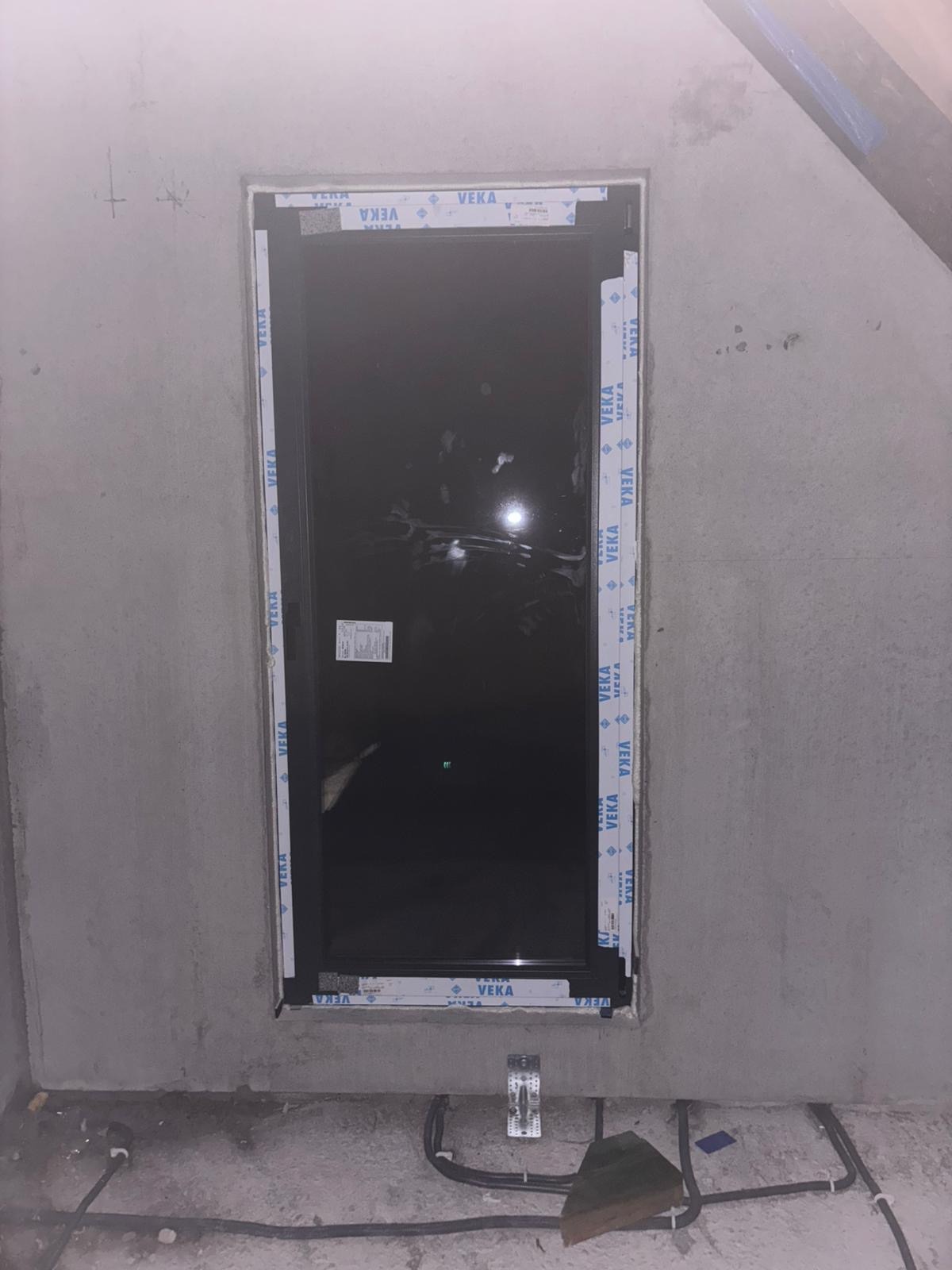 Czarne okno PCV marki VEKA w surowym otoczeniu, widoczne przewody instalacji elektrycznej przy podłodze. Etap montażu okna w trakcie prac wykończeniowych.