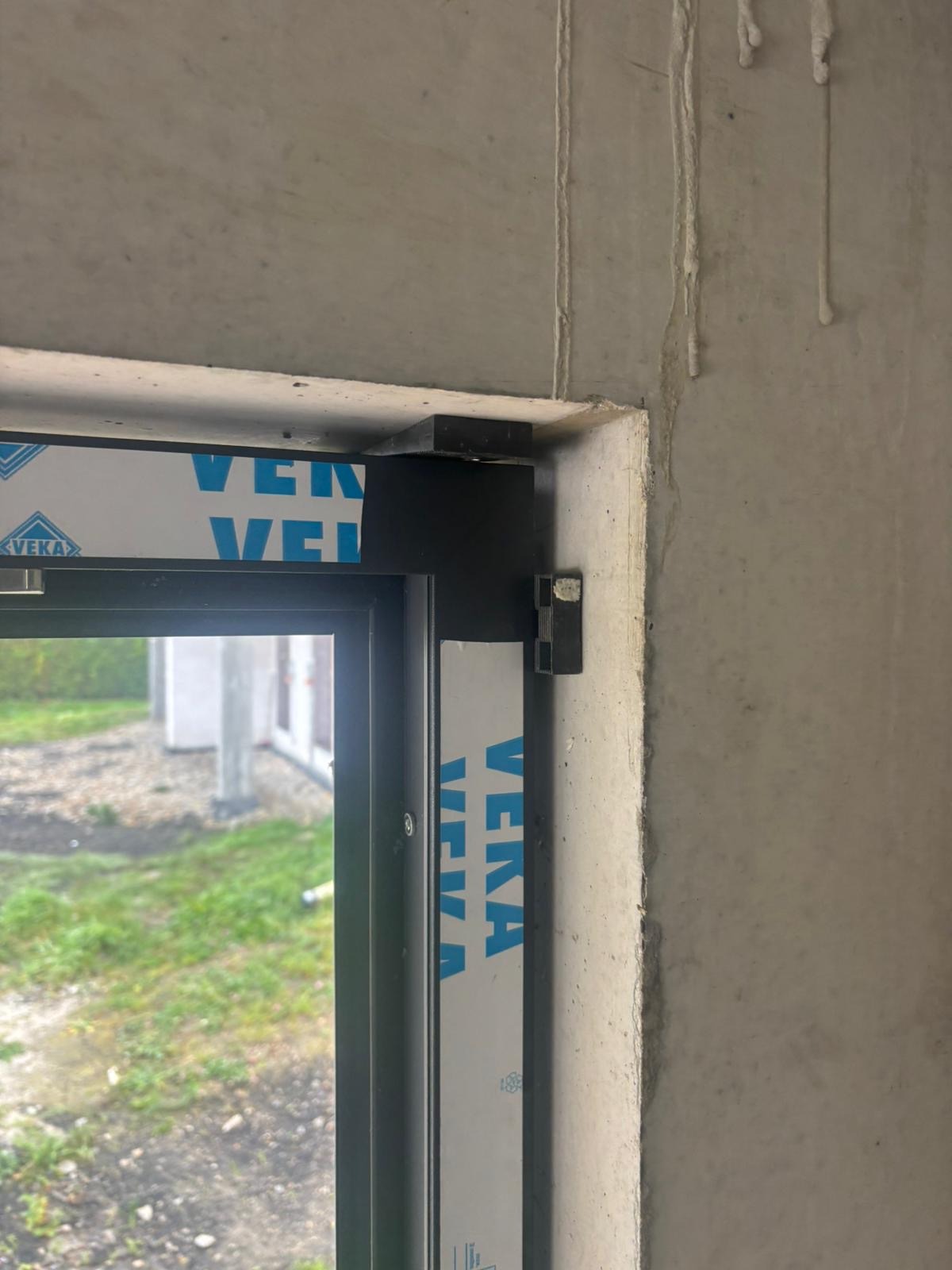 Fragment czarnej ramy okiennej z widocznym profilem marki Veka, osadzonej w surowym, betonowym otworze okiennym. Widoczne ślady po montażu.