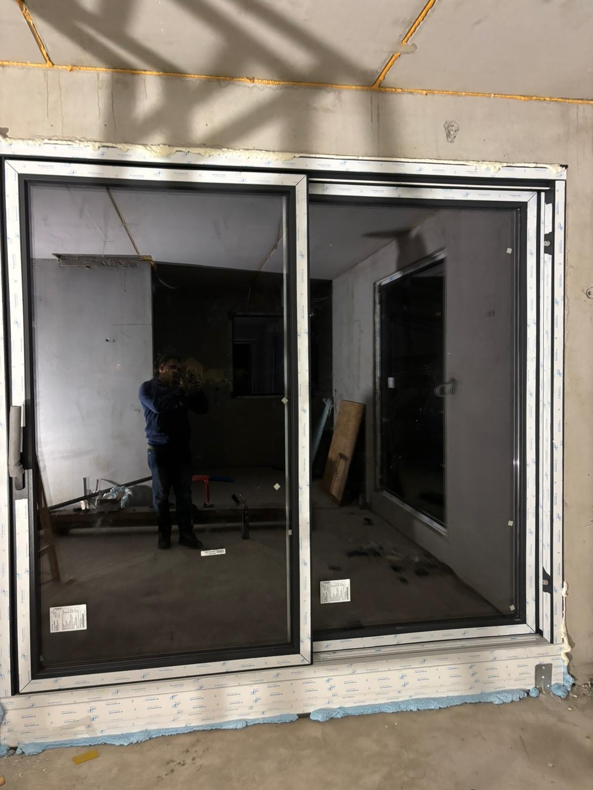 Zamontowane, czarne drzwi przesuwne z widocznym odbiciem osoby w szybie, osadzone w surowym otoczeniu budowlanym z widoczną pianką montażową.