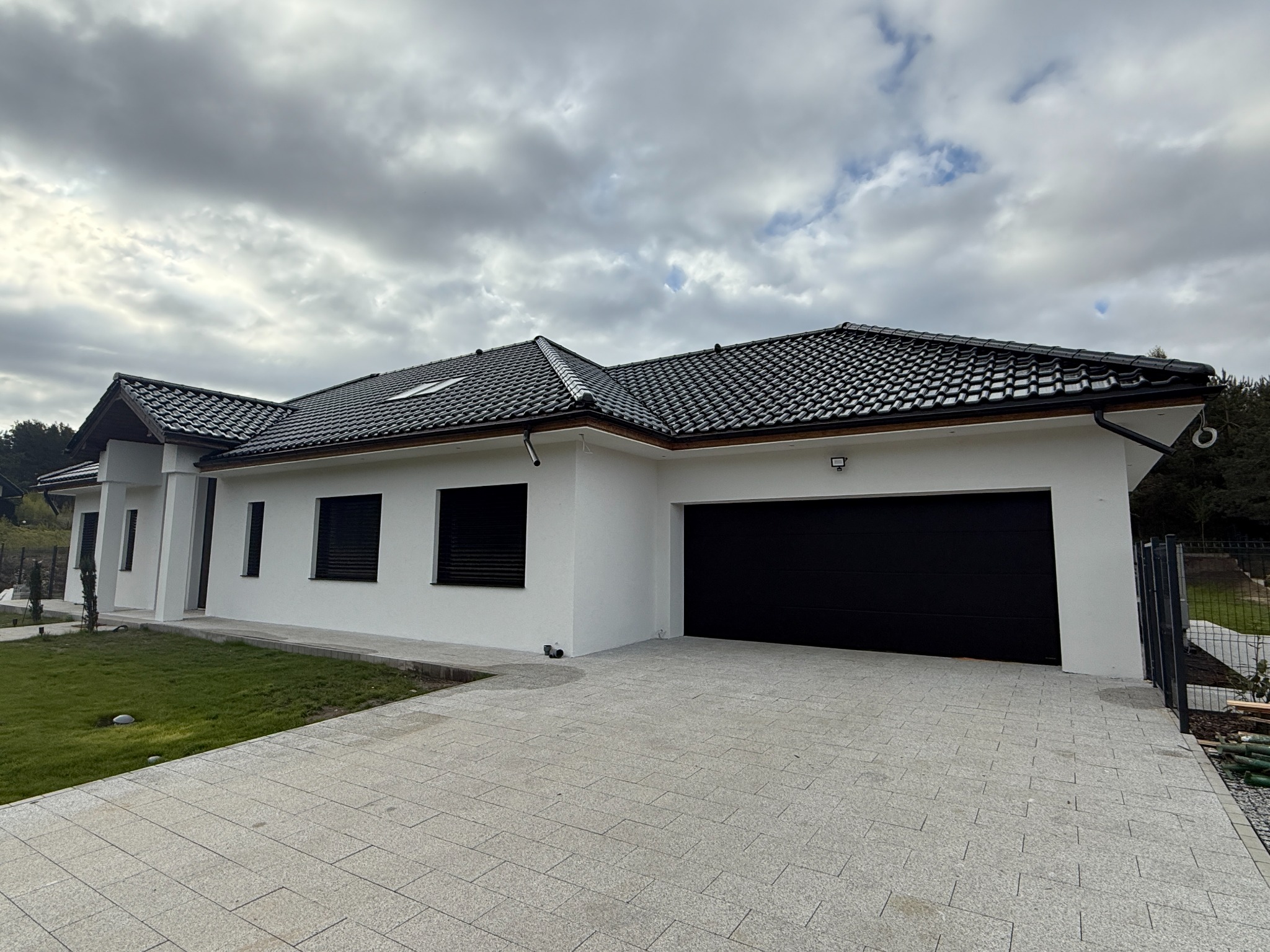 Nowoczesny dom jednorodzinny z czarną bramą garażową i oknami z roletami zewnętrznymi. Jasna elewacja, ciemna dachówka, kostka brukowa na podjeździe. Pochmurne niebo.