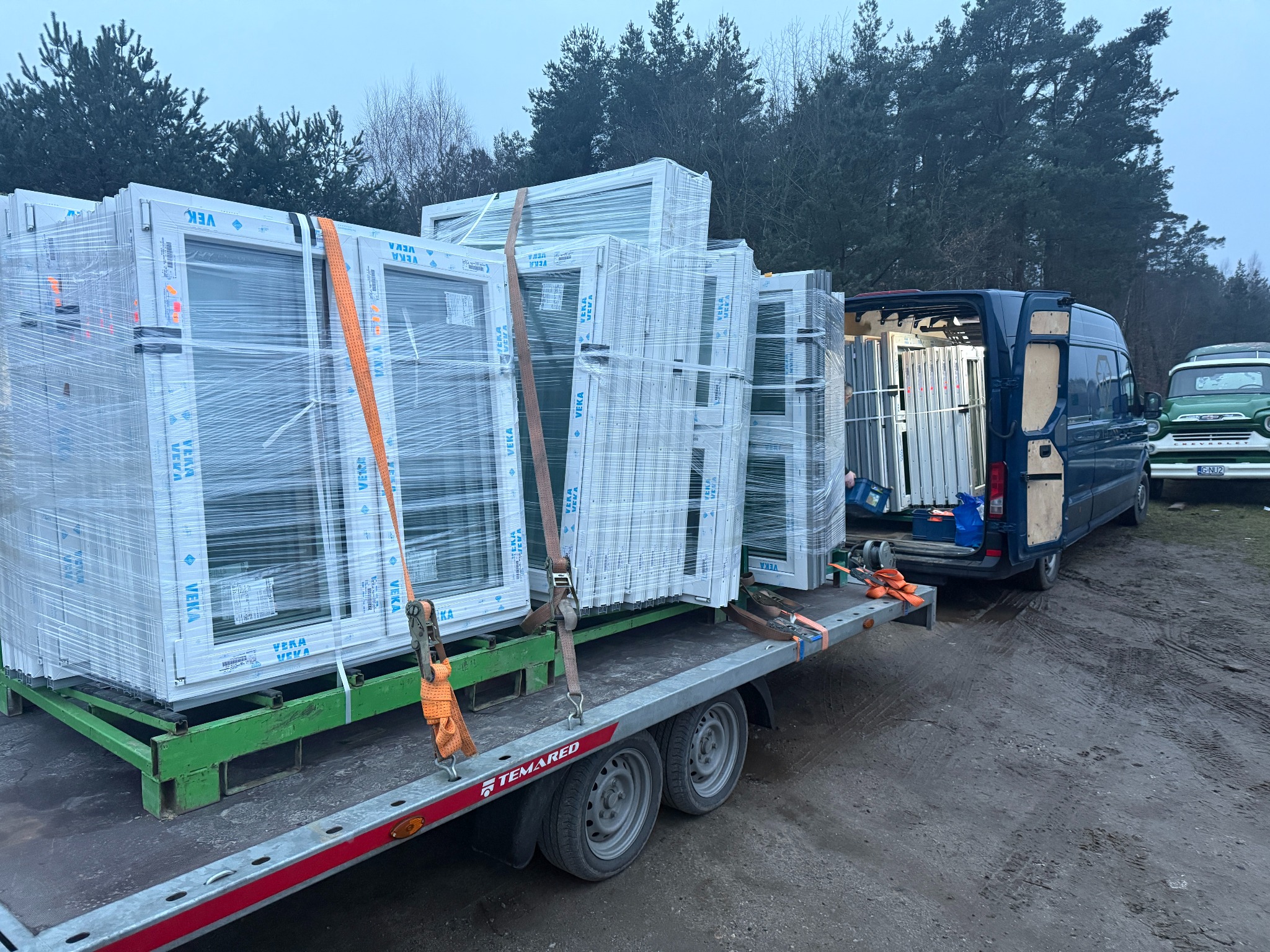 Transport okien PCV marki VEKA na przyczepie TEMAREd, zabezpieczonych pasami, z dostawczym busem w tle i zielonym Chevroletem na uboczu.