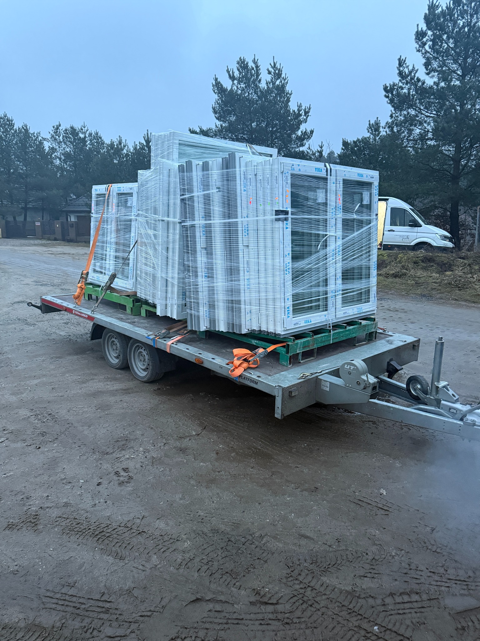 Transport okien PCV na przyczepie, zabezpieczonych folią i pasami. Widoczny las w tle i biały bus. Szare, pochmurne niebo.
