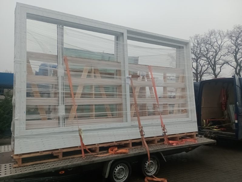 Duże okno PCV, zabezpieczone folią i pasami, na przyczepie transportowej. Widoczny fragment samochodu dostawczego i drzewa w tle. Przygotowanie do montażu.